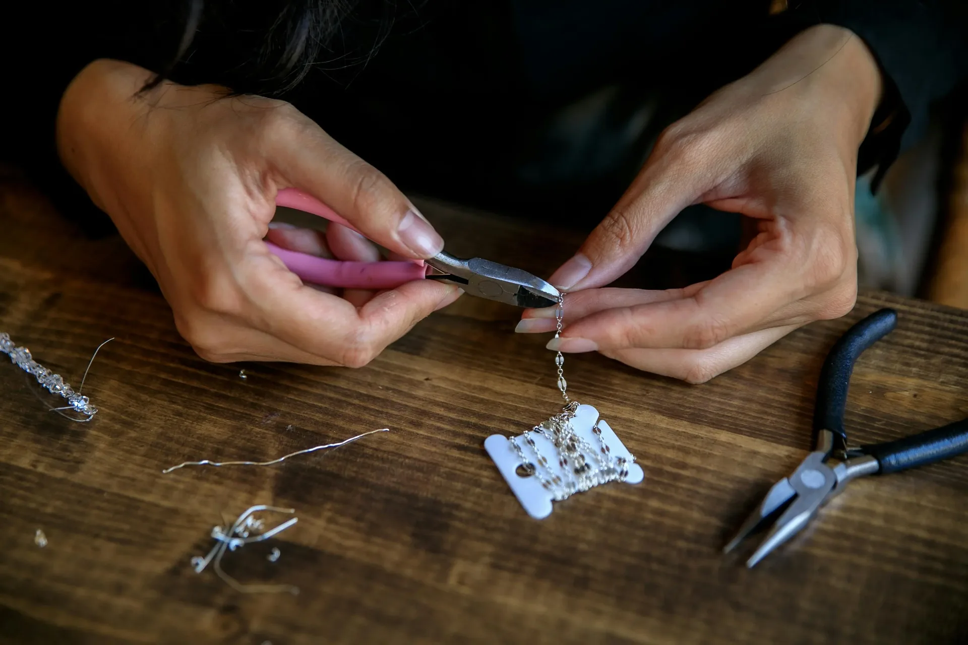 Artisan's hands using pliers to assemble a delicate silver chain on a wooden workbench surrounded by jewelry-making tools