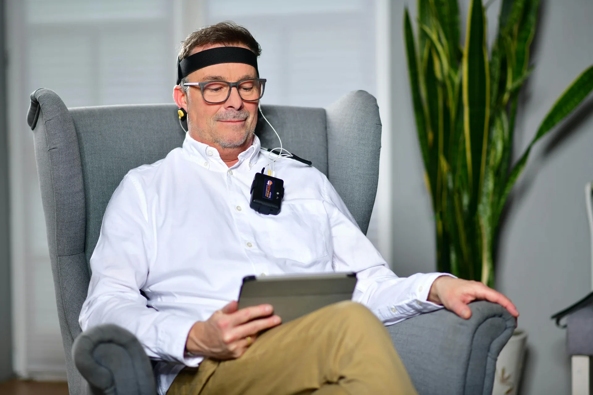 Middle-aged man relaxing in an armchair wearing a neurofeedback meditation headband and earbuds while using a tablet