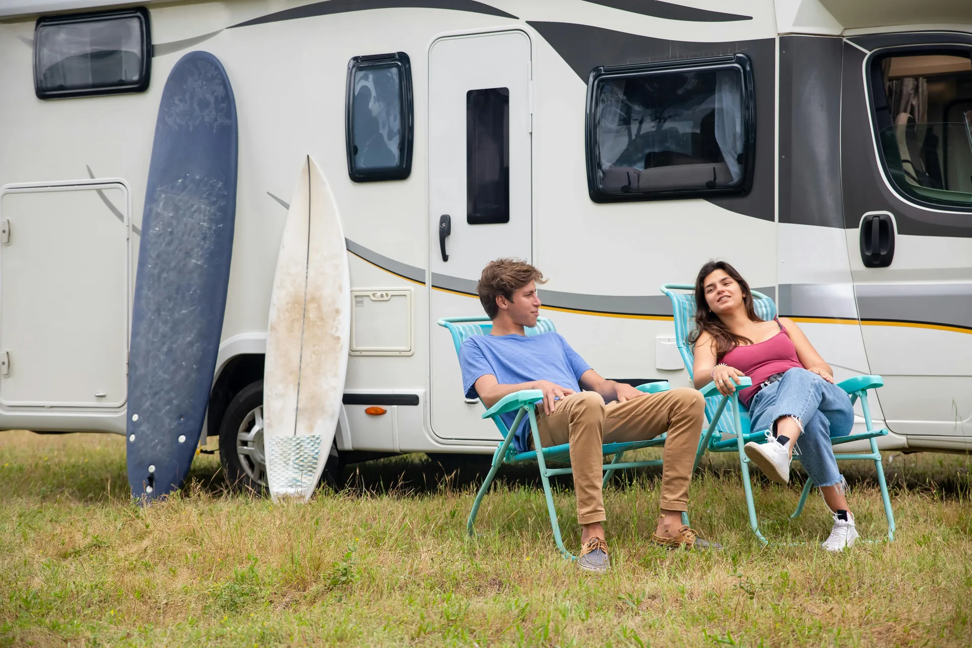 Young couple relaxing in camp chairs outside a parked RV with a surfboard leaning against the side
