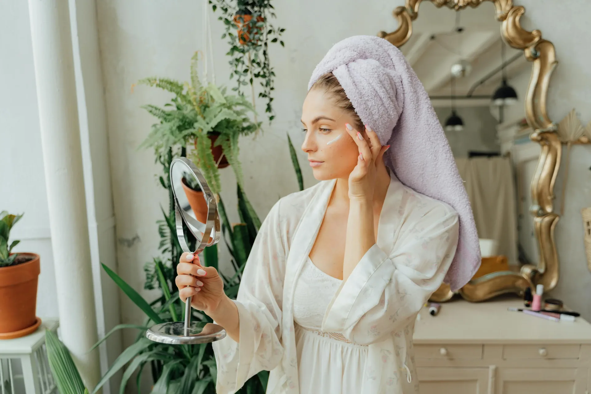 Woman with a lavender hair towel examining her face in a gold ornate hand mirror during her natural skincare routine